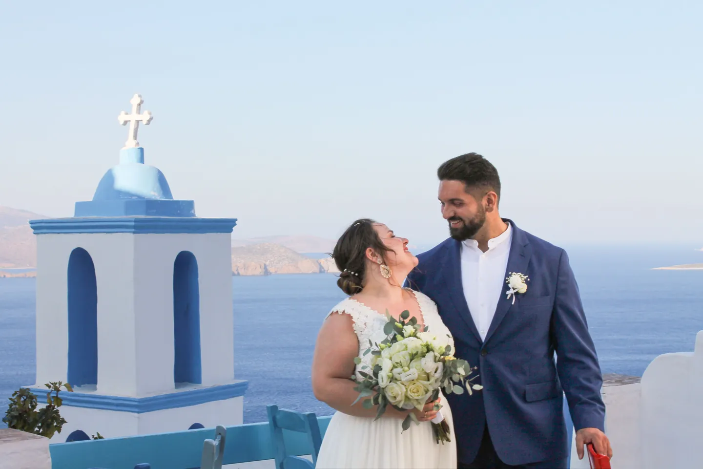 A bride and groom smile at each other on a terrace above the sea, next to a white chapel with a blue dome