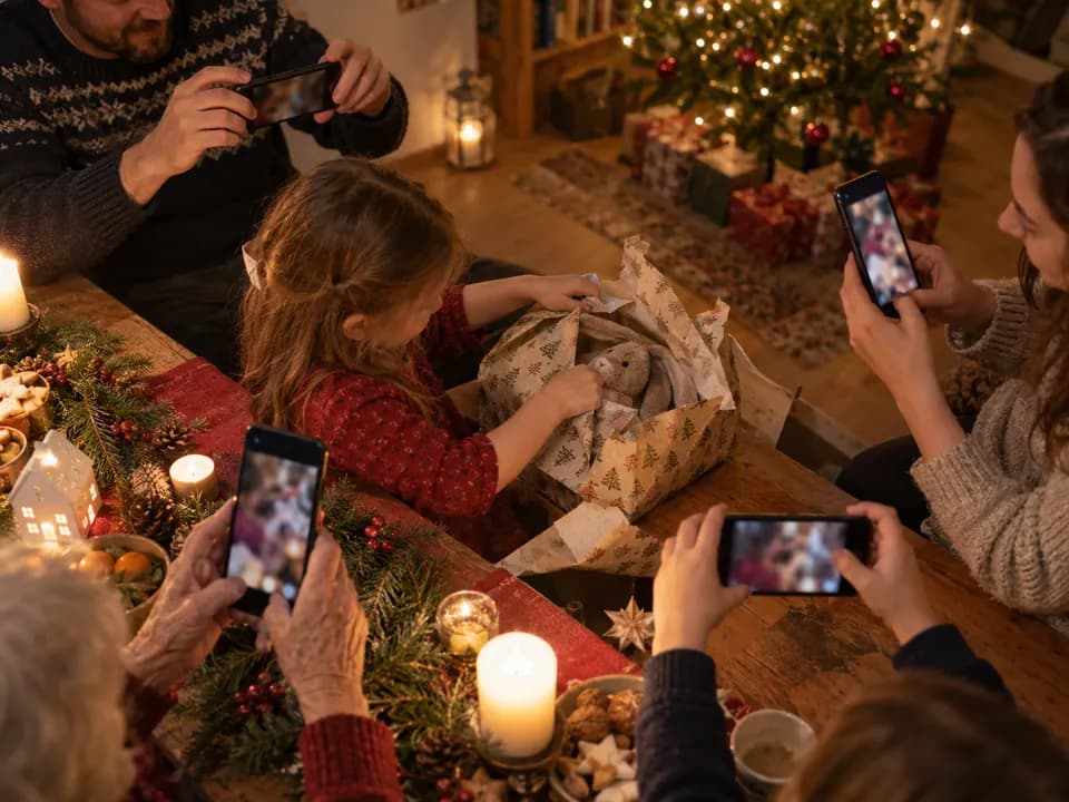 Mehrere Hände verschiedener Generationen halten Handys über den Weihnachtstisch und fotografieren denselben Moment aus verschiedenen Richtungen